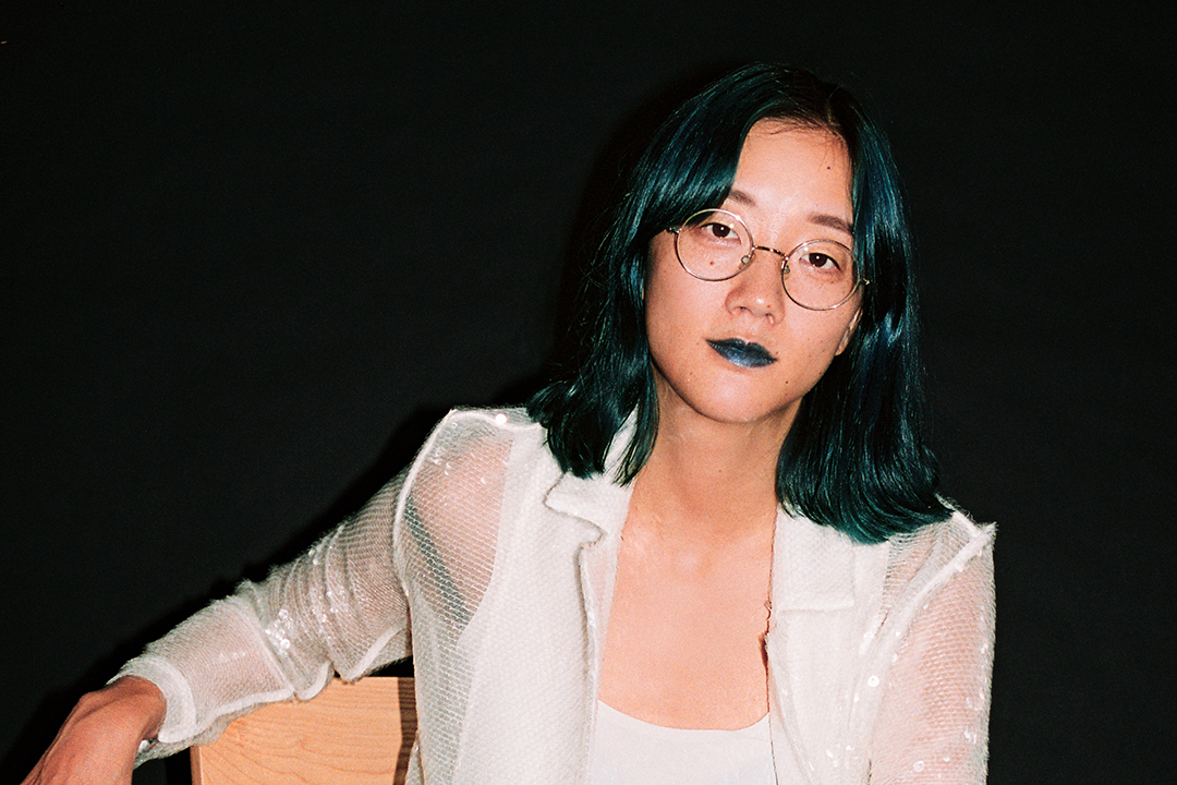 Christine Sun Kim, her hair dyed a dark teal with matching lipstick, sits on a colorful wooden chair in front of a black background. She is wearing a sheer jacket over a white tank top and black leather pants. Her gaze is intense but slightly unfocused, as though her mind were on something else while observing the viewer.
